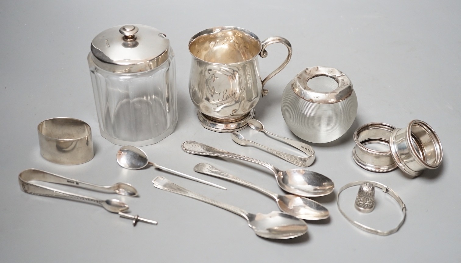 Sundry small silver including a mounted glass match tidy(a.f.), a silver christening mug(a.f.), mounted glass preserve jar, three napkin rings, six items of flatware, thimble T-bar and a sterling bangle.